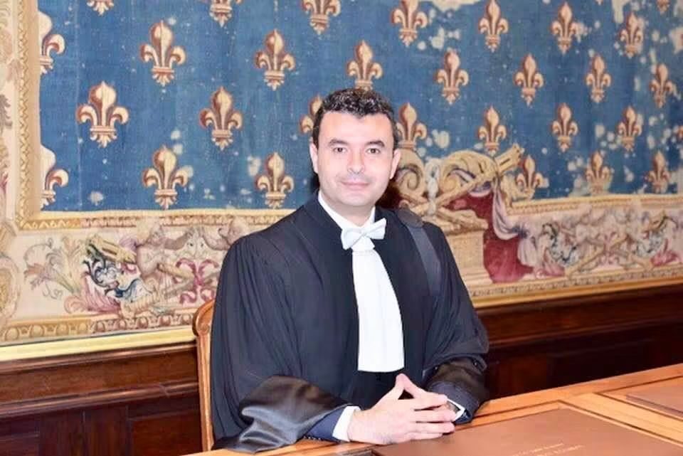 Maître Abdelatif Laamrani, en robe d'avocat, assis au Palais de justice de Paris devant la tapisserie fleurdelisée